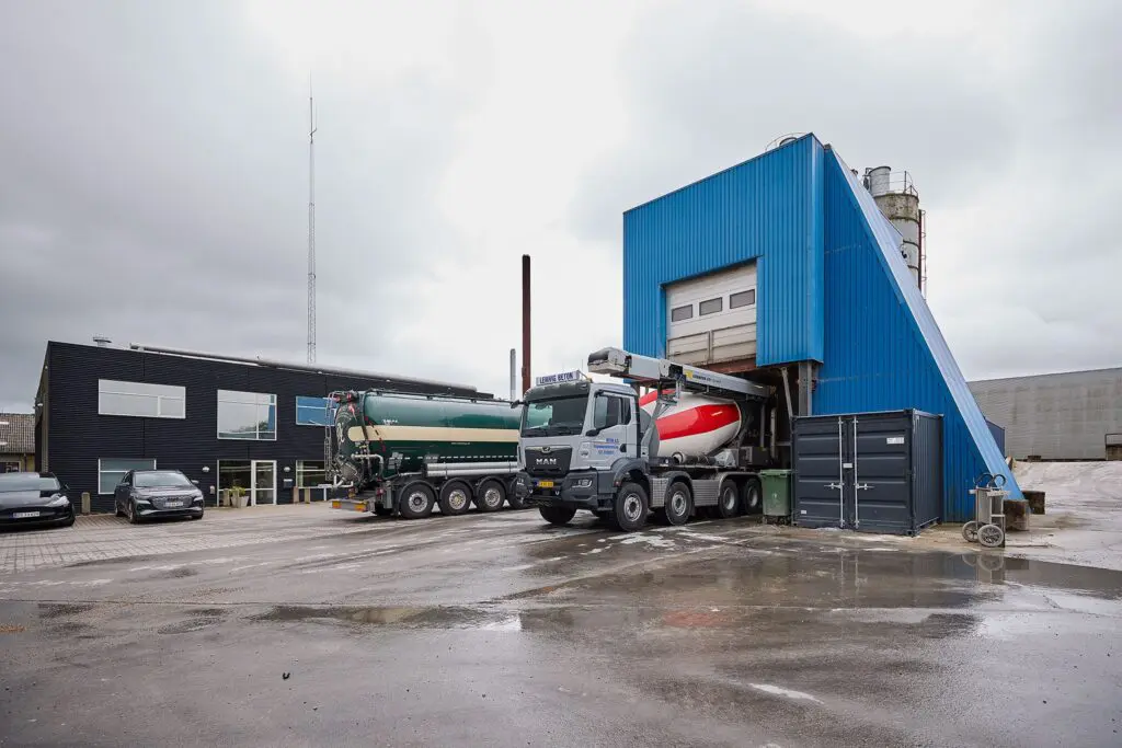 En MAN betonbil fra Lemvig Beton bliver fyldt op ved et blåt betontårn ved siden af en stor grøn tankbil.