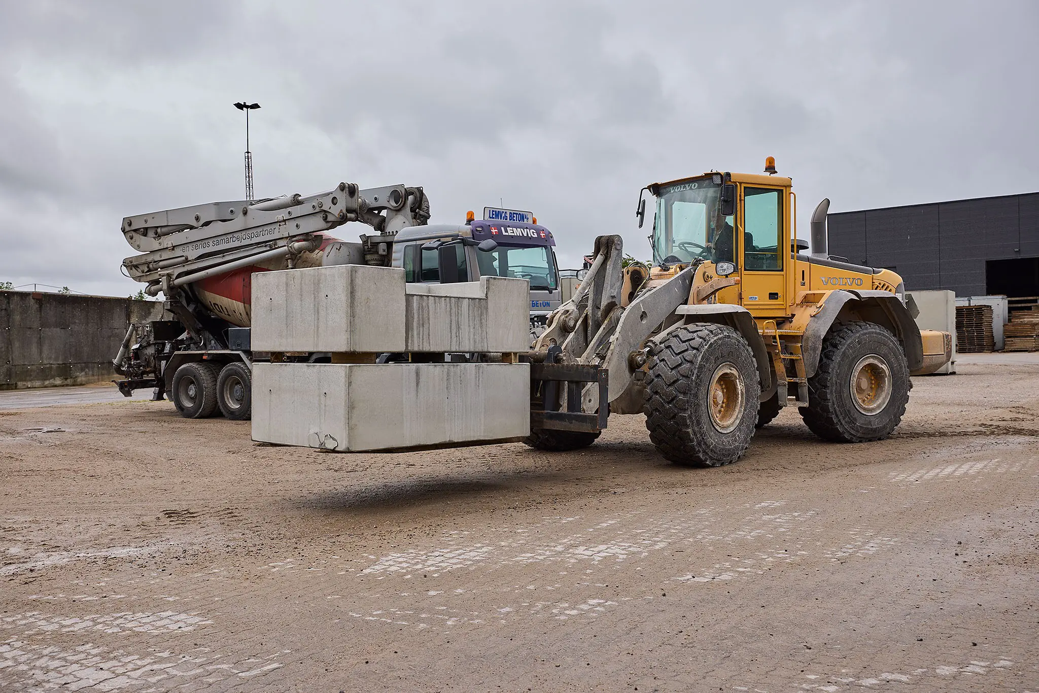 En gul Volvo gummiged løfter store betonblokke på en plads med en betonbil i baggrunden.