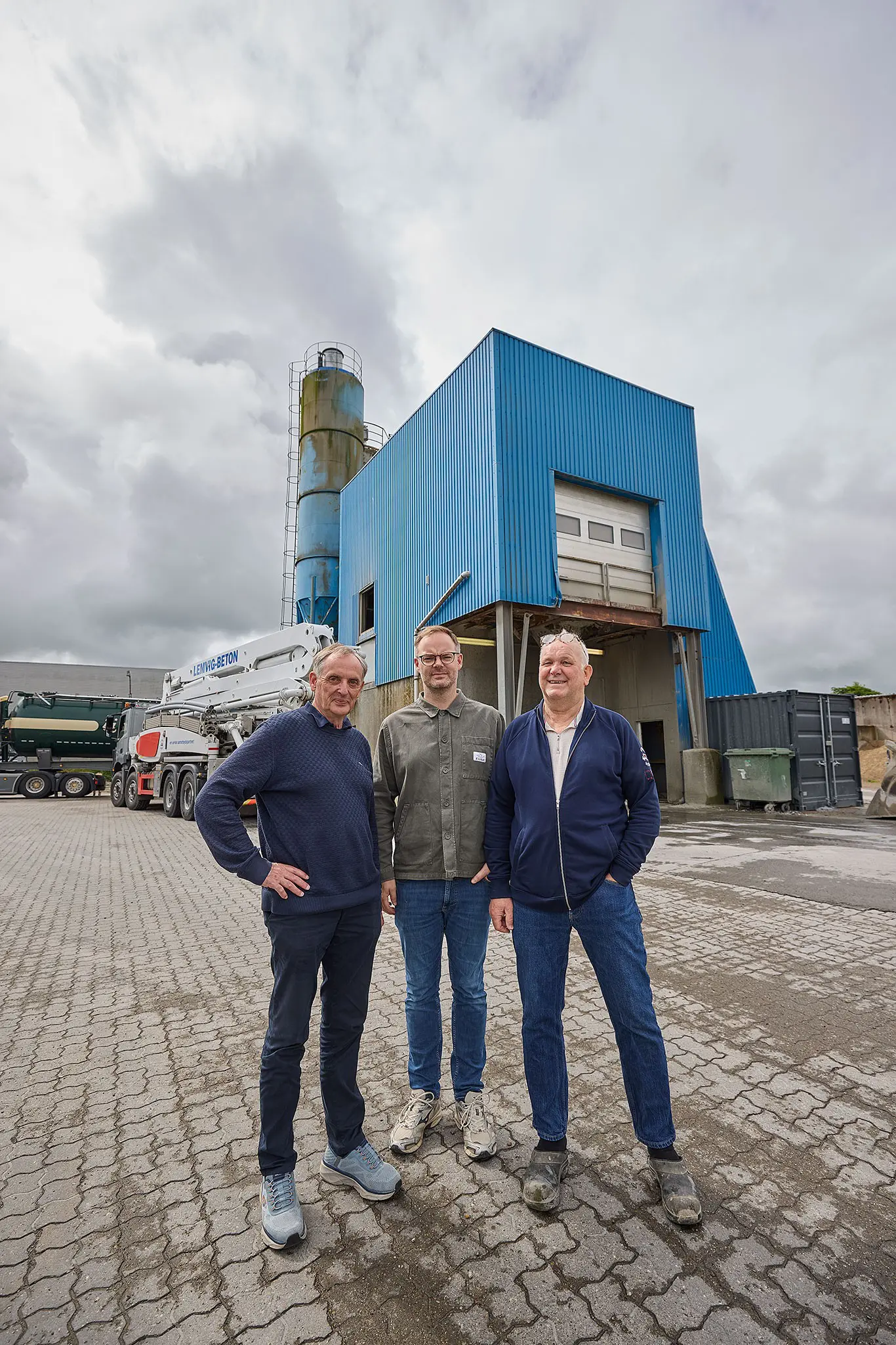 Tre mænd står foran Lemvig Betons blå industribygning med en betonpumpebil i baggrunden.